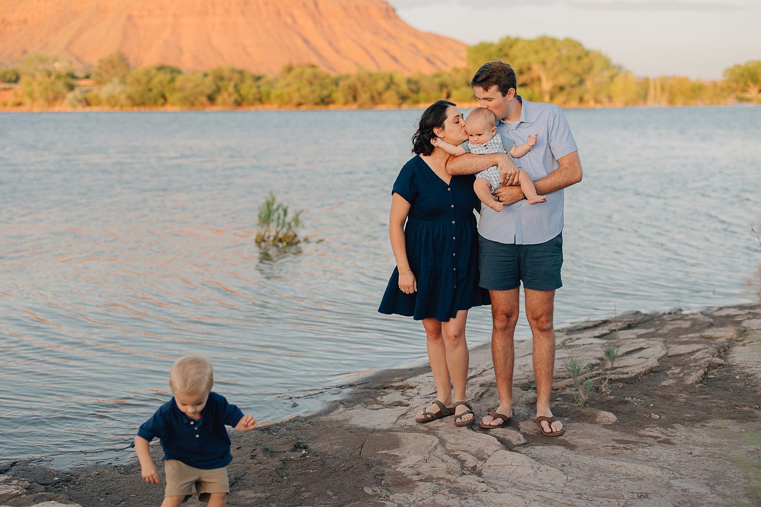 Family of four enjoying golden hour by a scenic lake with red rock mountains, parents kissing baby while toddler plays nearby.