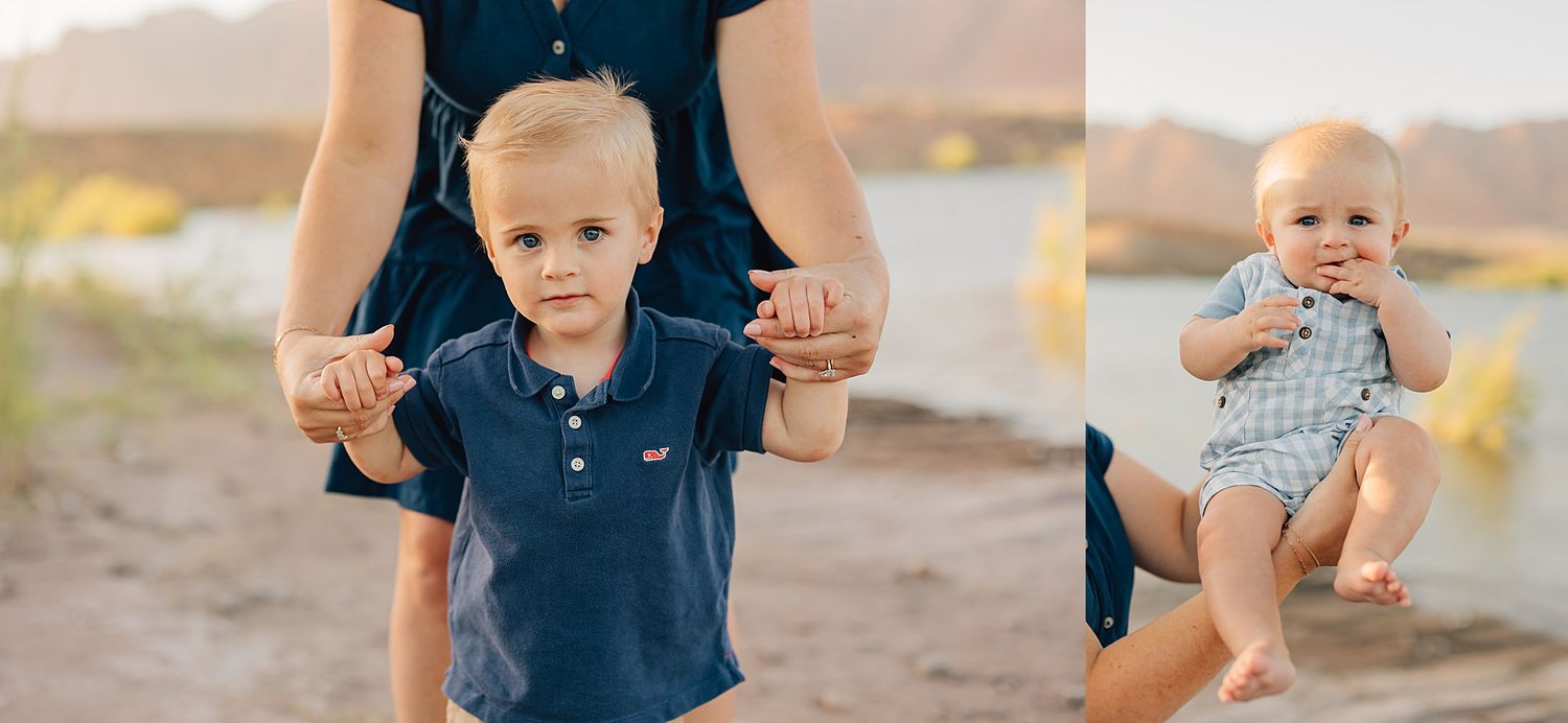 A toddler boy holds his mother's hands on a sandy beach, beside a baby boy held by an adult in a blue dress outdoors.