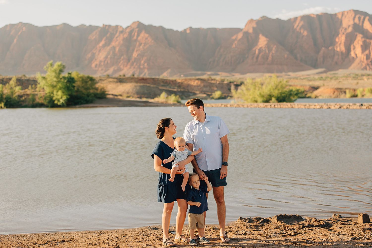 Family of four posing by a scenic lake with red rock mountains in the background on a sunny day.