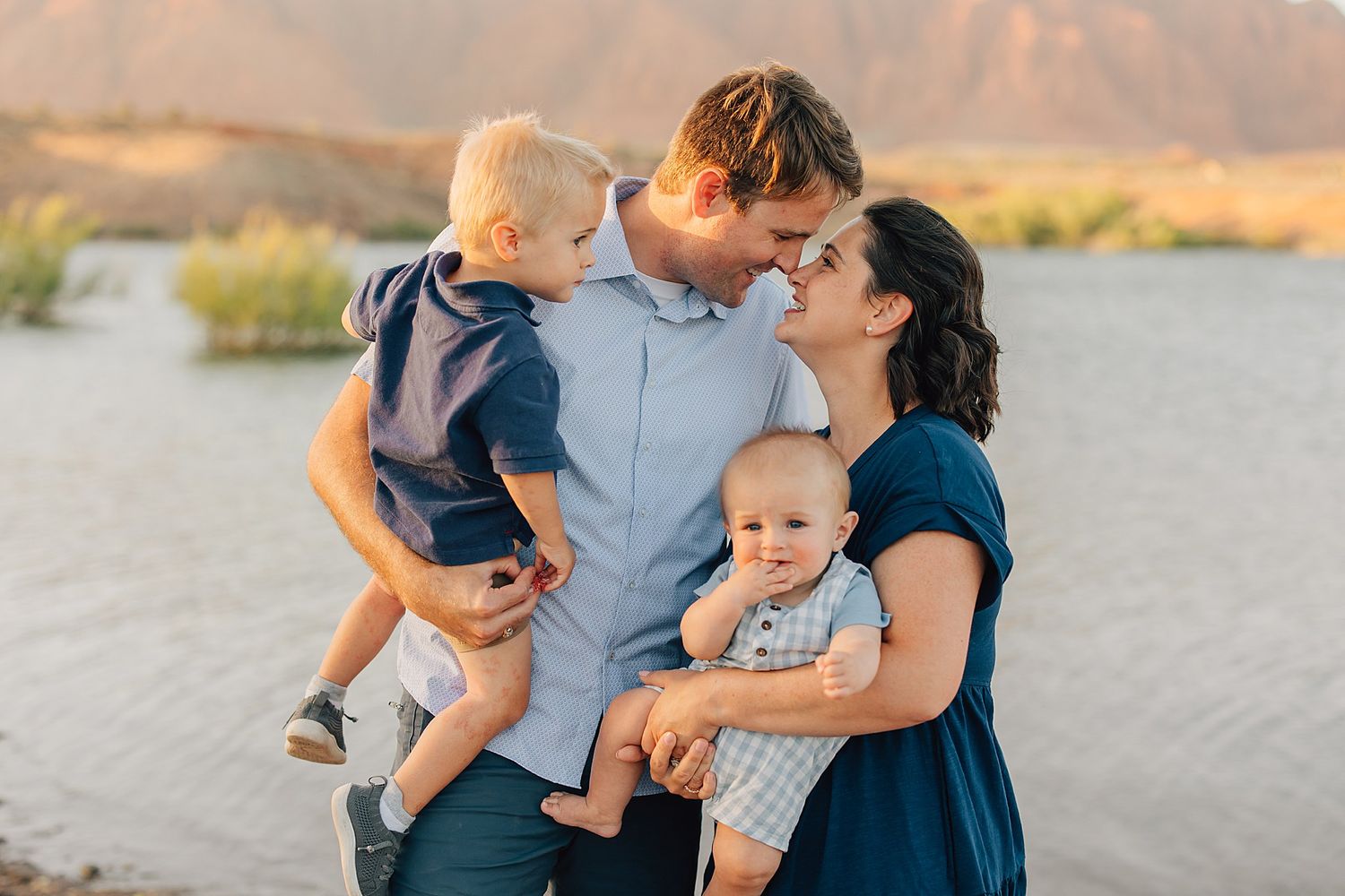 A happy young family with two toddlers shares a tender moment outdoors near a lake at golden hour.