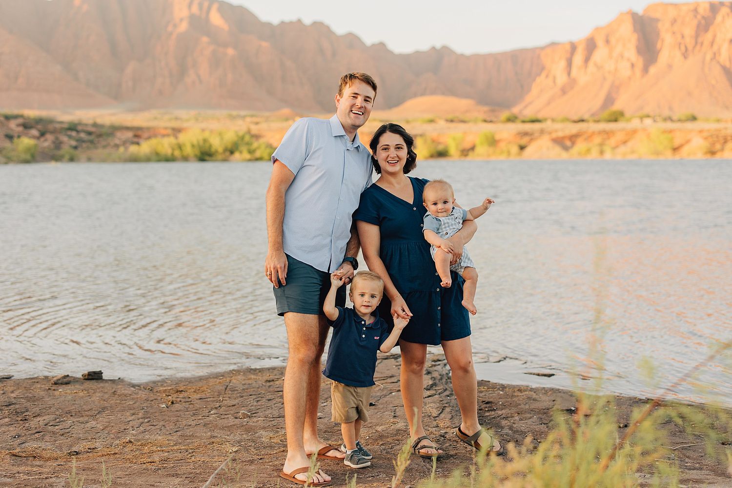 Family of four posing by a river with red rock mountains at golden hour in a desert landscape.