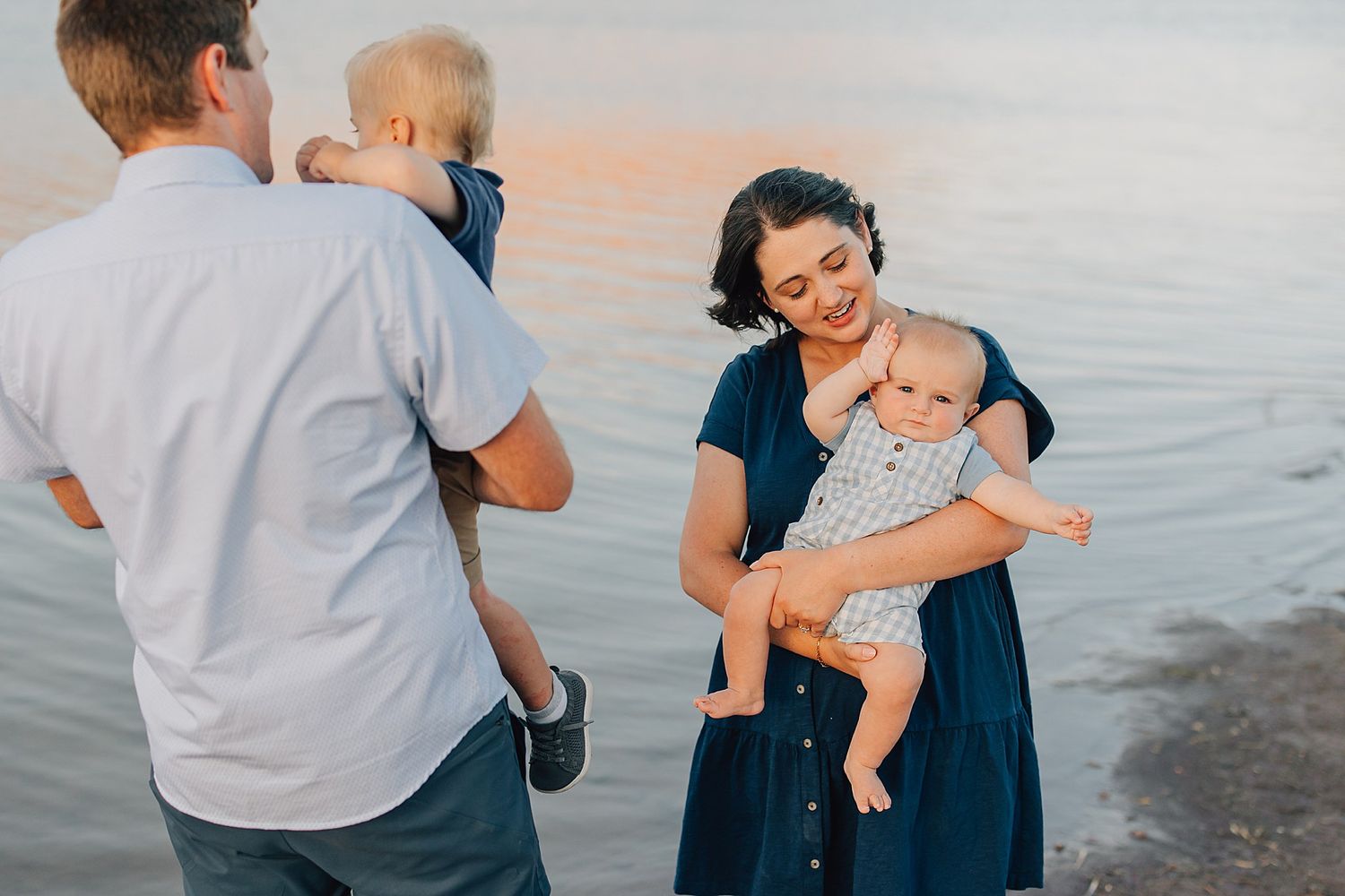 Family with two babies posing near calm water at sunset, mother in navy dress holding infant, father carrying toddler.