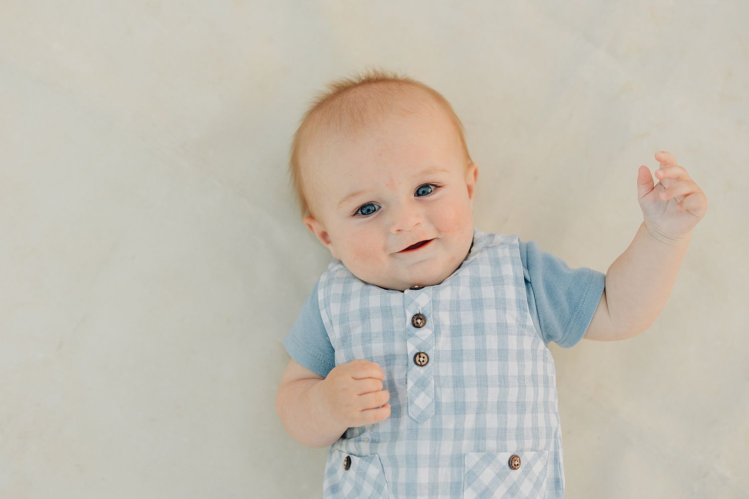 Adorable smiling baby in blue gingham outfit lying on white blanket, looking up with bright eyes.