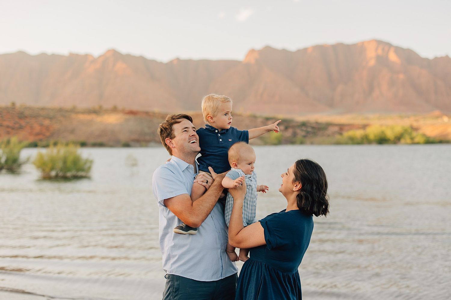 Family of four posing by a lake with mountain backdrop at golden hour, father holding toddler, mother cradling baby.