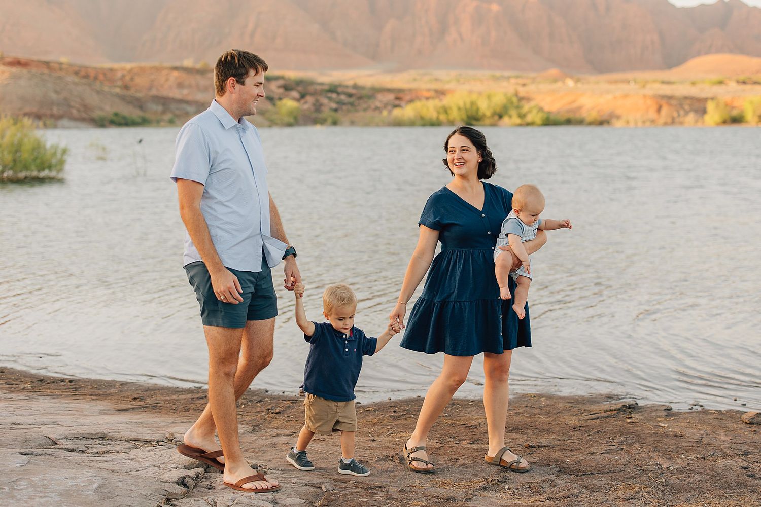 Family of four walking hand-in-hand along a scenic lakeside with red rock mountains at golden hour.