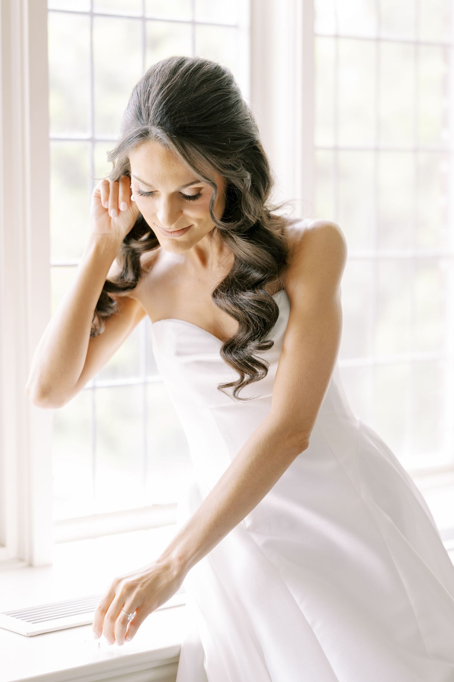 Elegant bride in strapless white gown sitting by a bright window, with flowing dark hair and a serene smile.