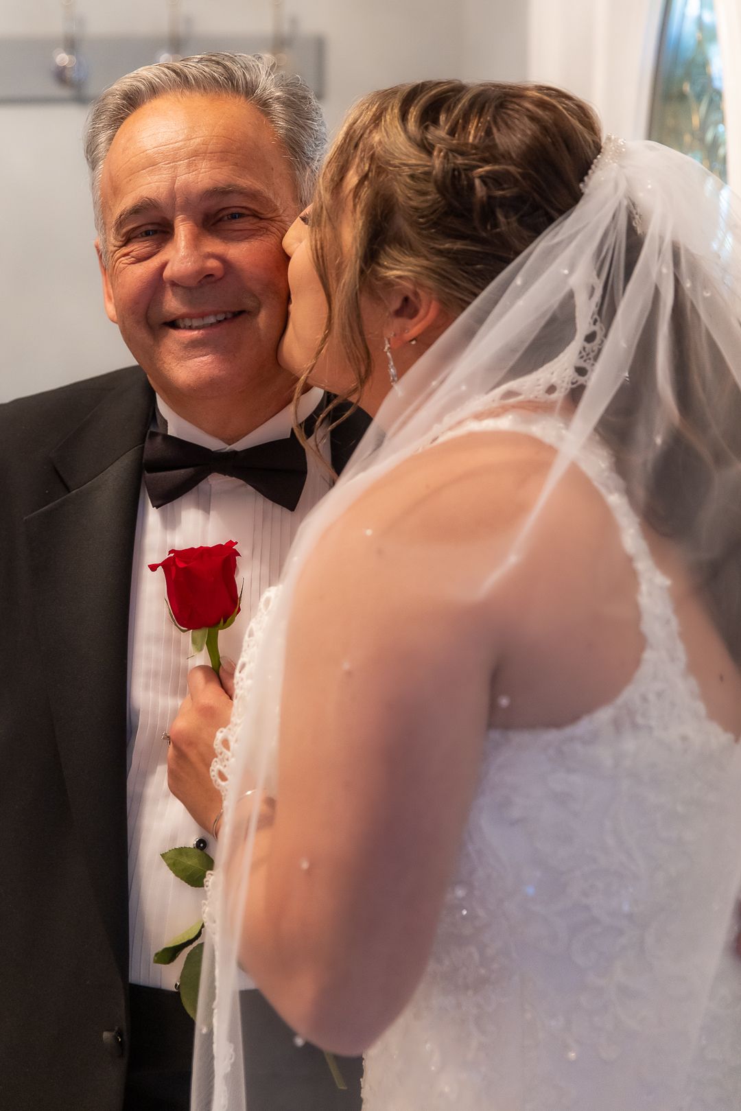 Bride in white gown and veil kisses older man in tuxedo holding a red rose at a wedding ceremony.