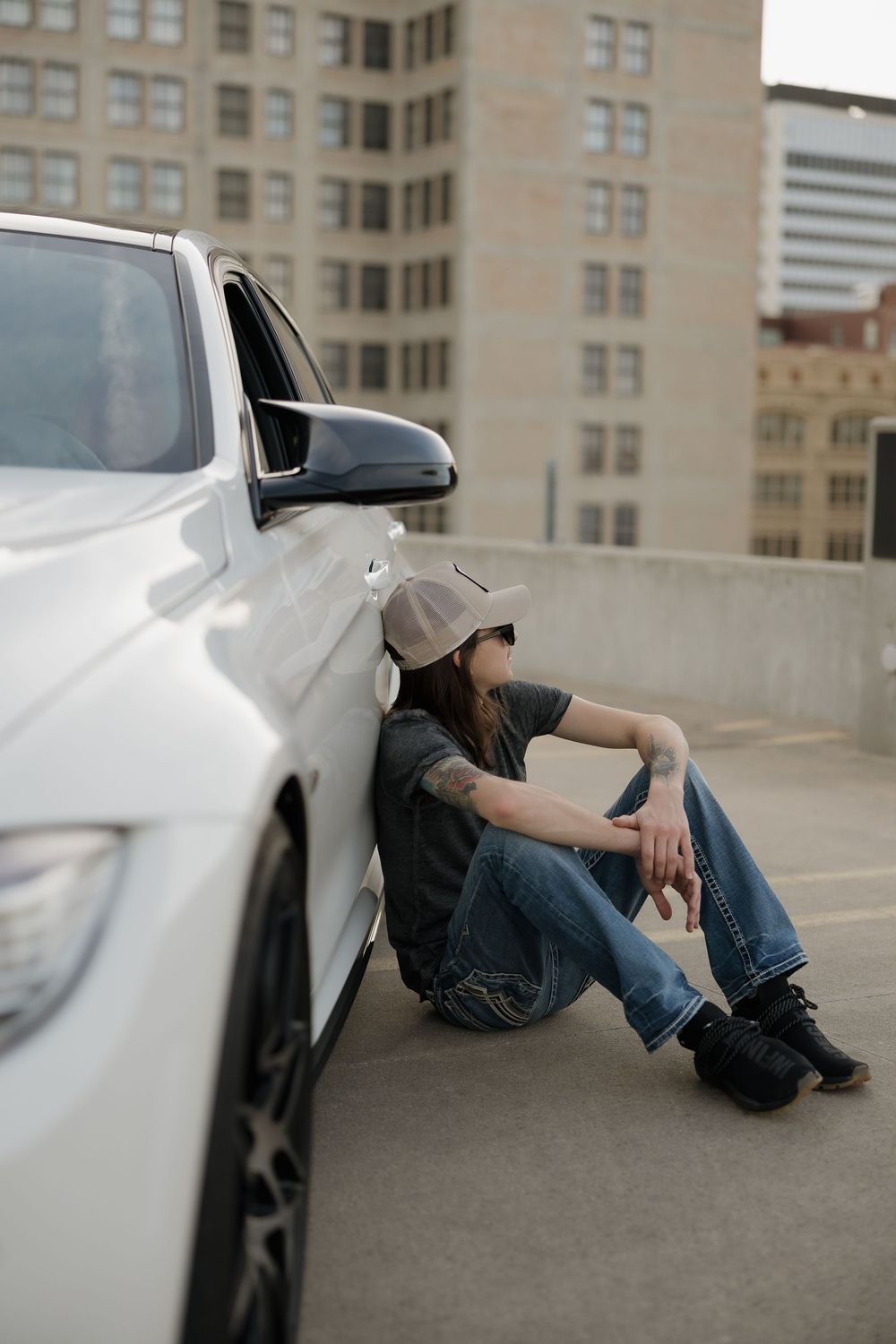 Person in cap and sunglasses sitting against white BMW on urban rooftop parking lot.