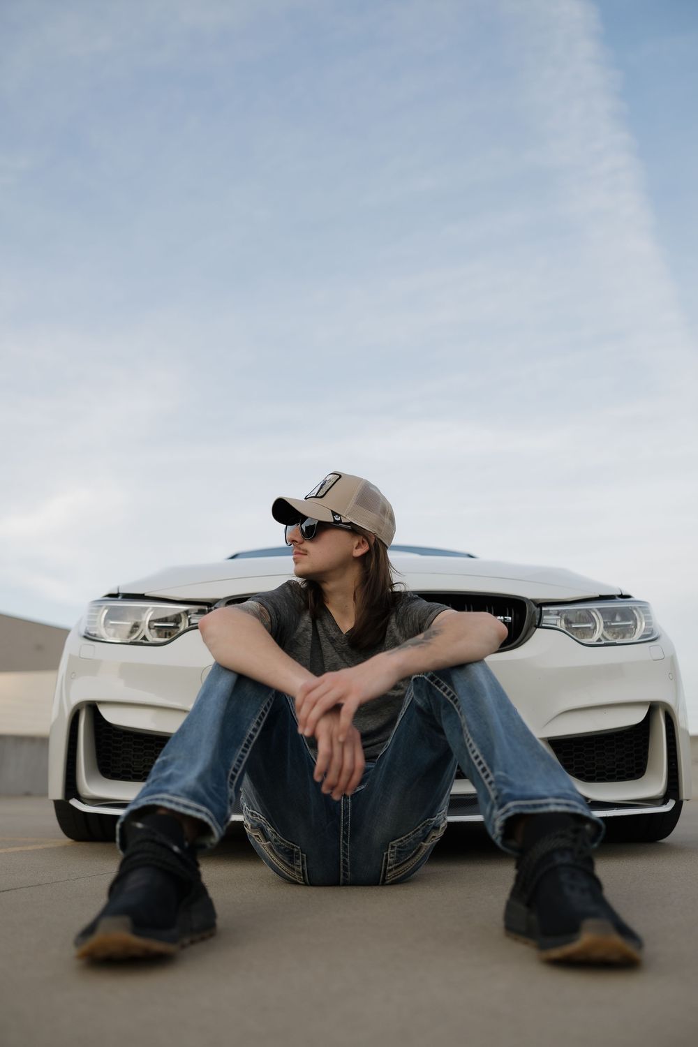 Stylish person in cap and sunglasses sitting in front of white BMW on rooftop parking lot.