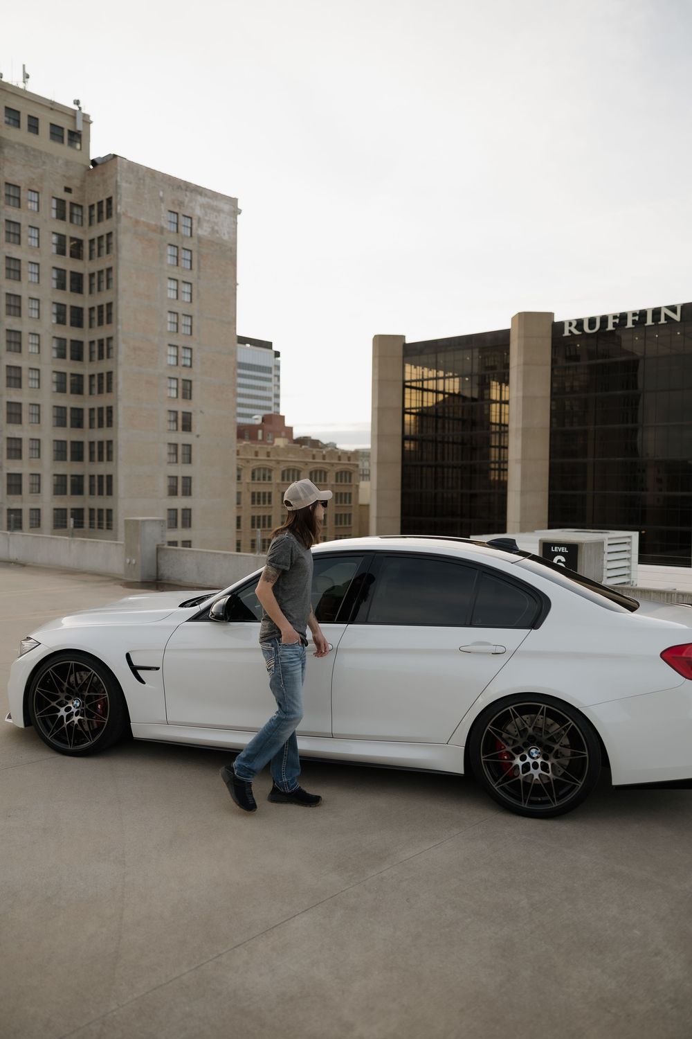Person in cap standing beside white BMW M3 on rooftop parking lot with city buildings.