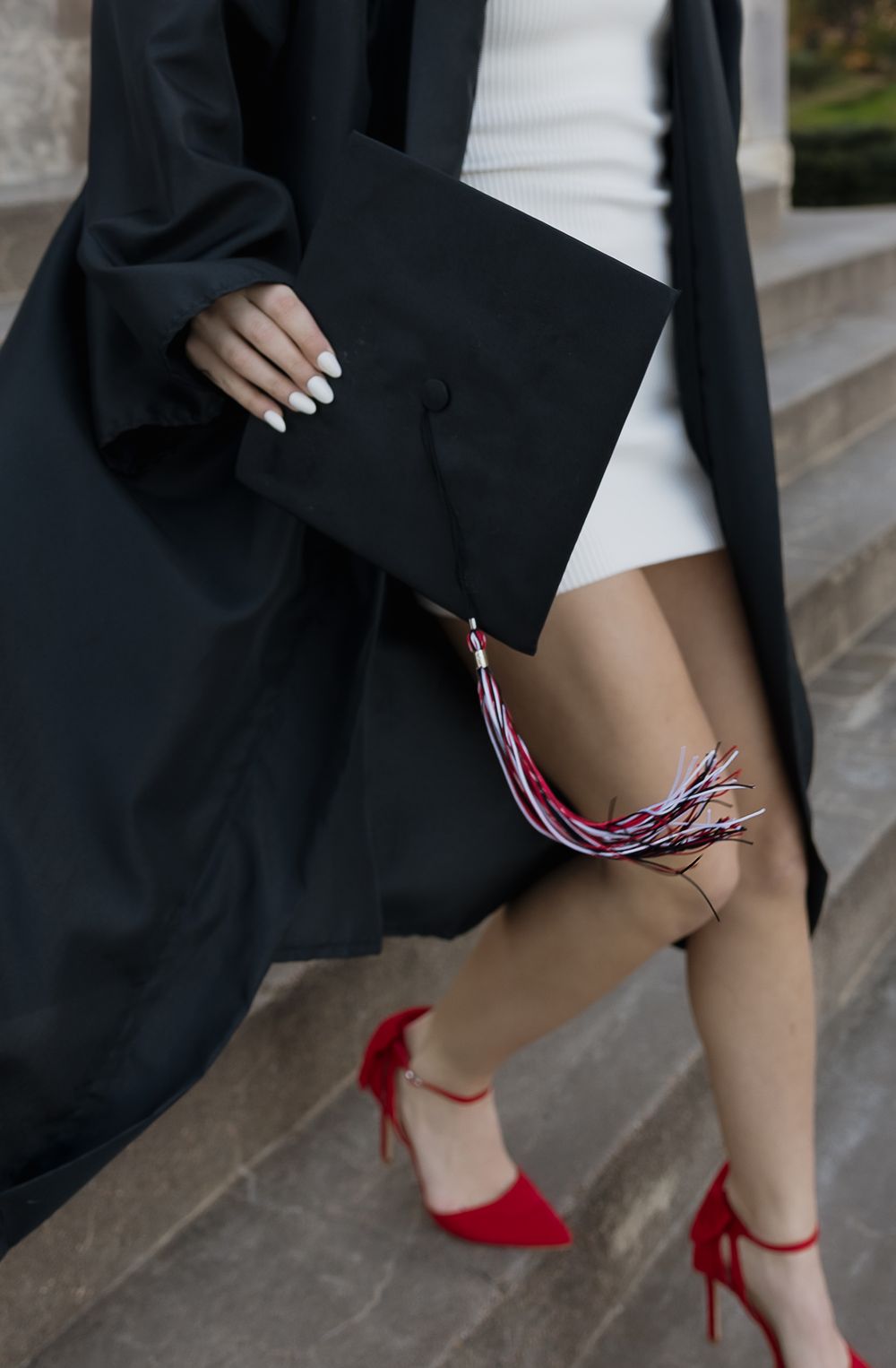 Graduate in black gown holding mortarboard cap with colorful tassel wearing red heels.