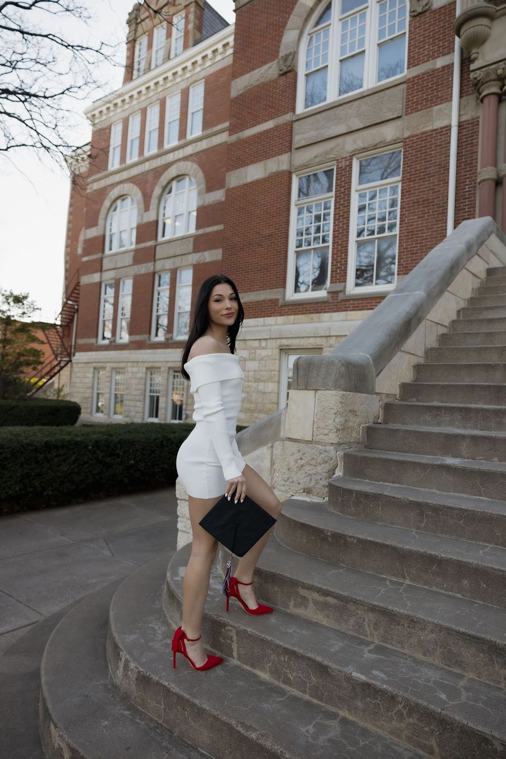 Young woman in white off-shoulder dress and red heels on university building steps.