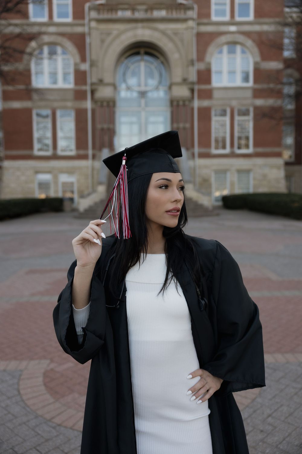 Female graduate in black cap and gown wearing white dress in front of brick university.