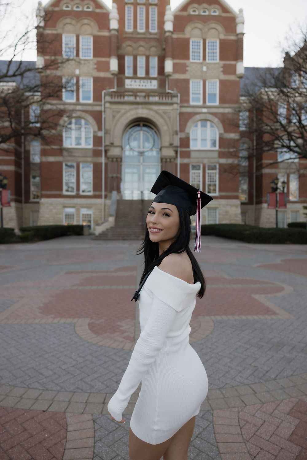 Smiling female graduate in white dress and black mortarboard cap on brick campus plaza.