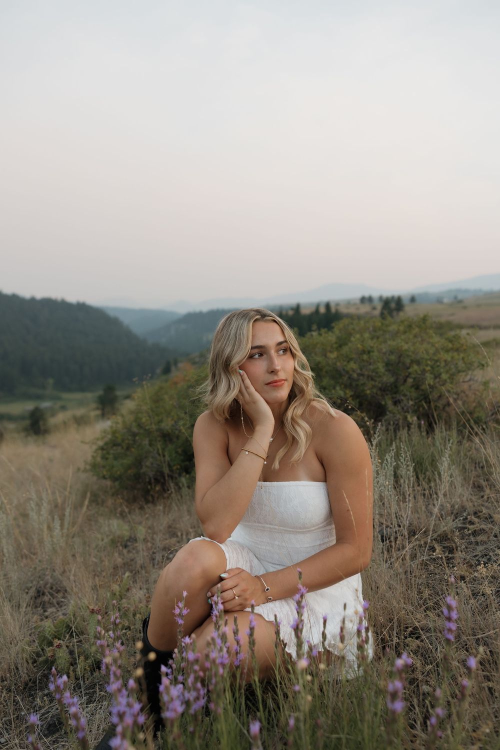 Young blonde woman in white strapless dress sitting in wildflower field with mountain valley backdrop at dusk.