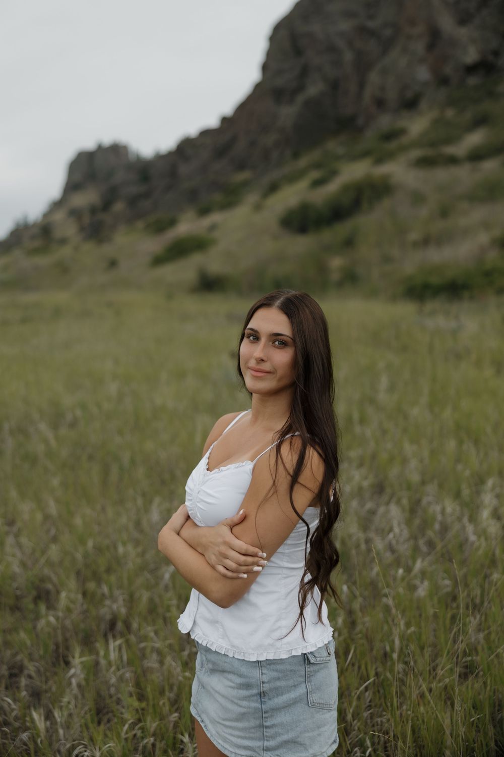 Young woman in white top and denim skirt posing in a green meadow with rocky hills in the background.
