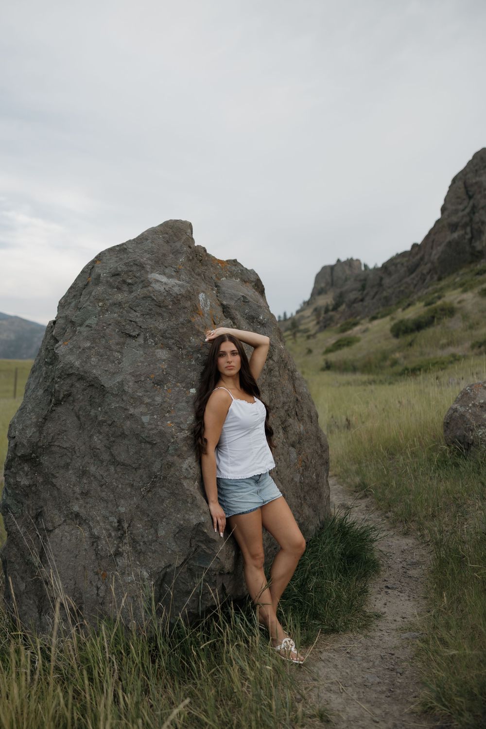 Young woman in white tank top and denim shorts leaning against large boulder in scenic mountain meadow.