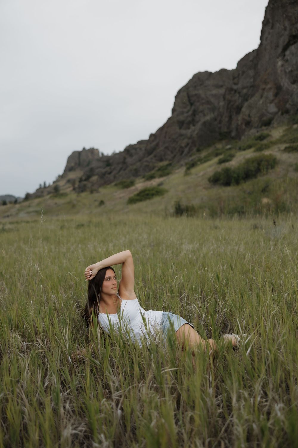 Woman in white dress reclines gracefully in tall green meadow grass with rocky mountain cliffs in the background.