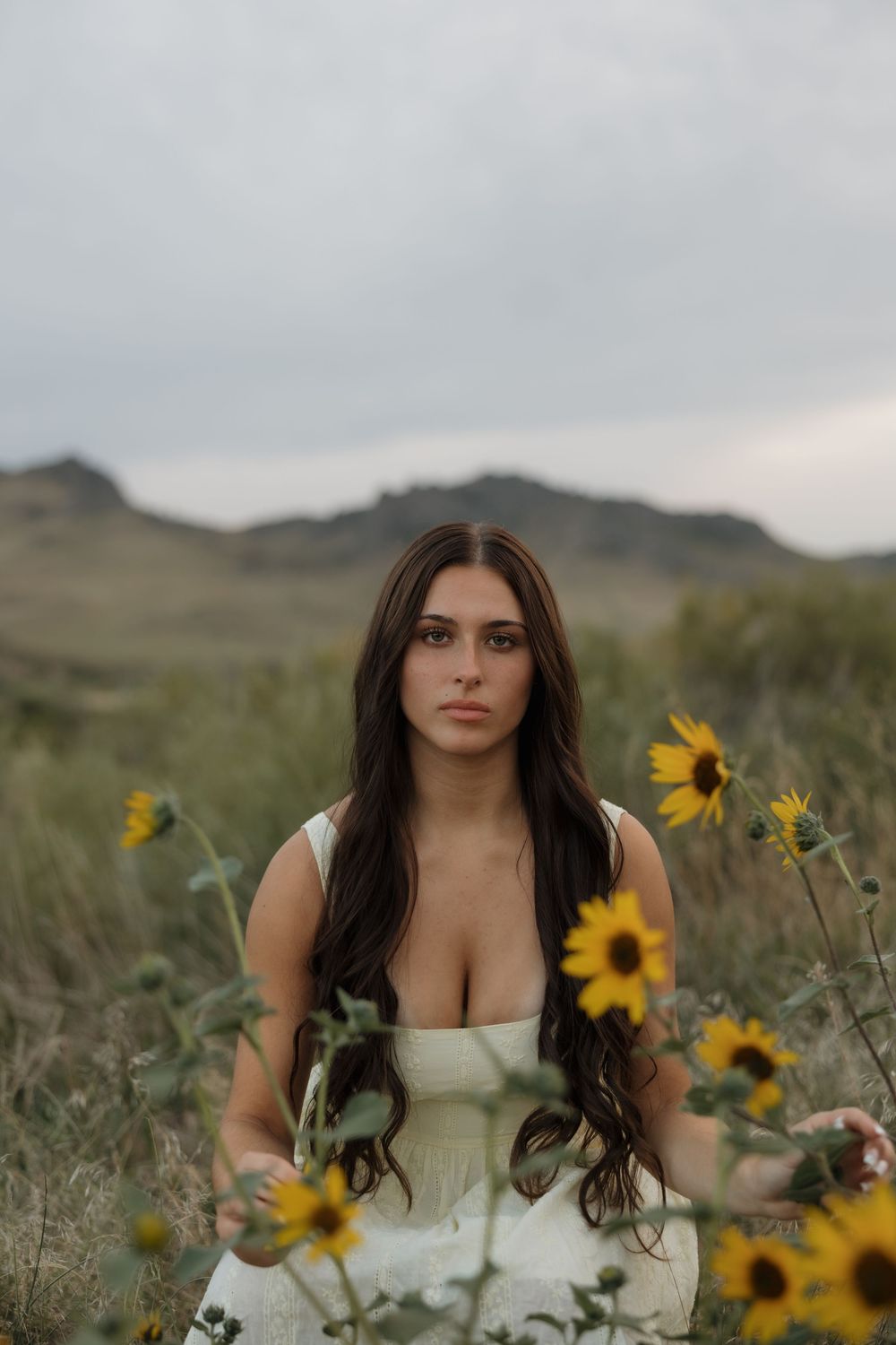 Young woman in white dress surrounded by sunflowers in a field with mountains in the background.