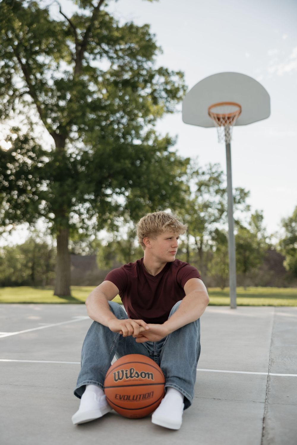 Young man sitting on outdoor basketball court leaning on Wilson Evolution basketball with hoop in background.