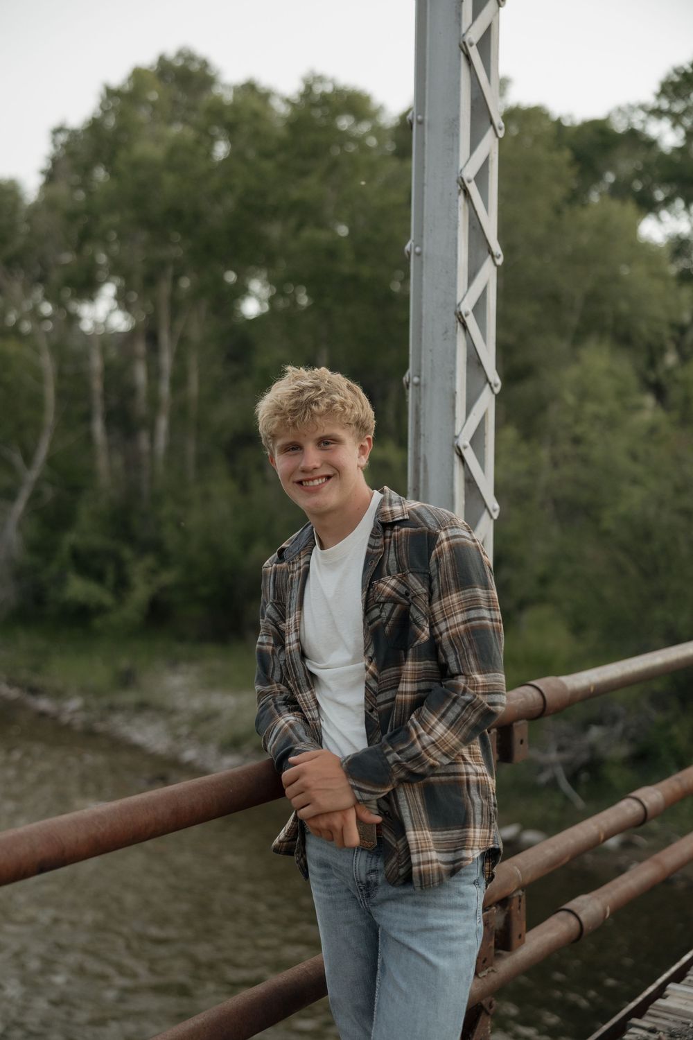 Smiling teenage boy in plaid shirt and jeans leaning on a rusty bridge railing with green trees behind.