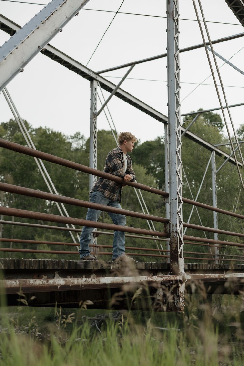 Young man in plaid shirt and jeans leaning on rusty railing of old metal bridge surrounded by trees.