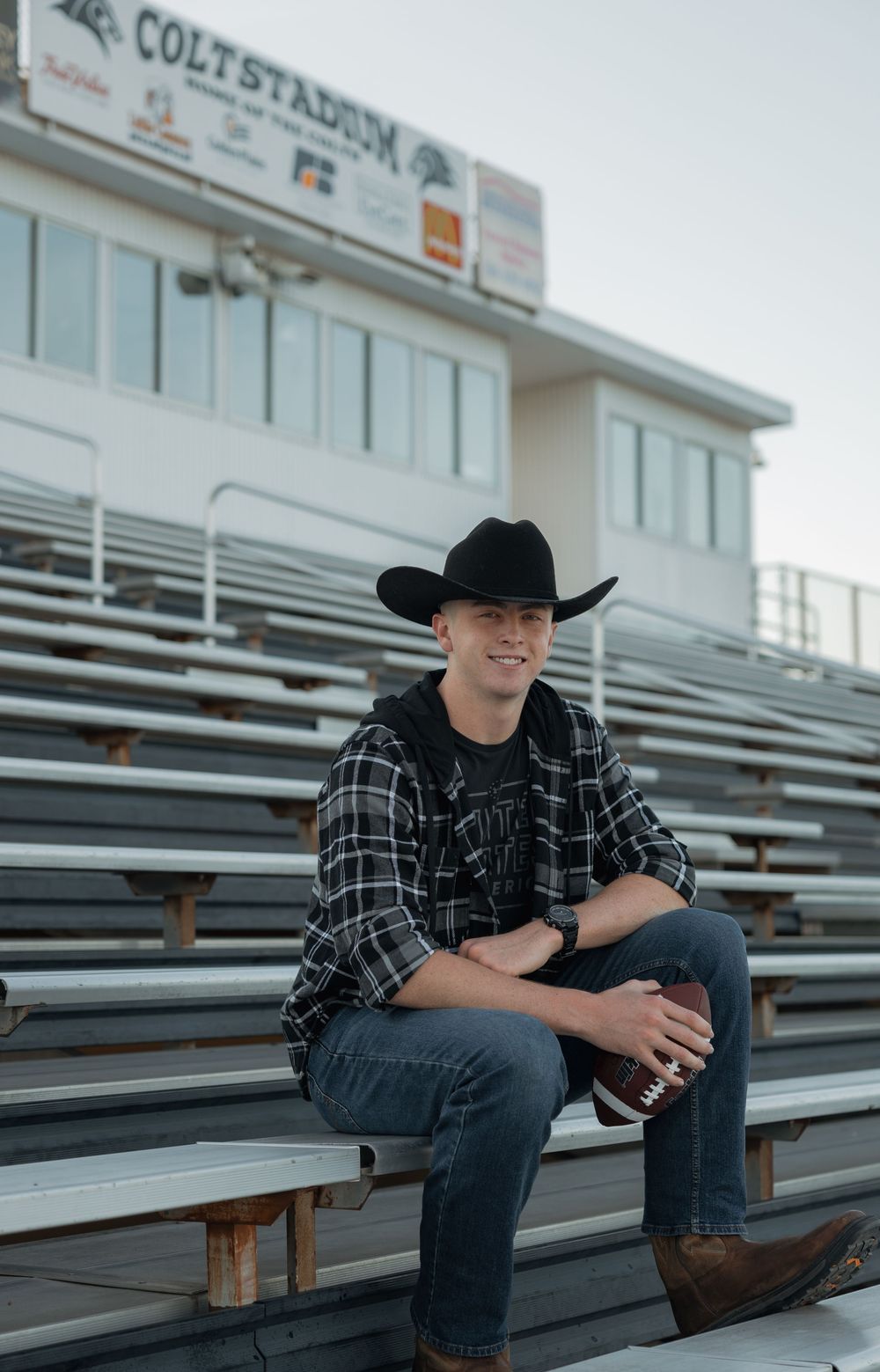 Young man in a black cowboy hat and plaid shirt sits in stadium bleachers holding a football.