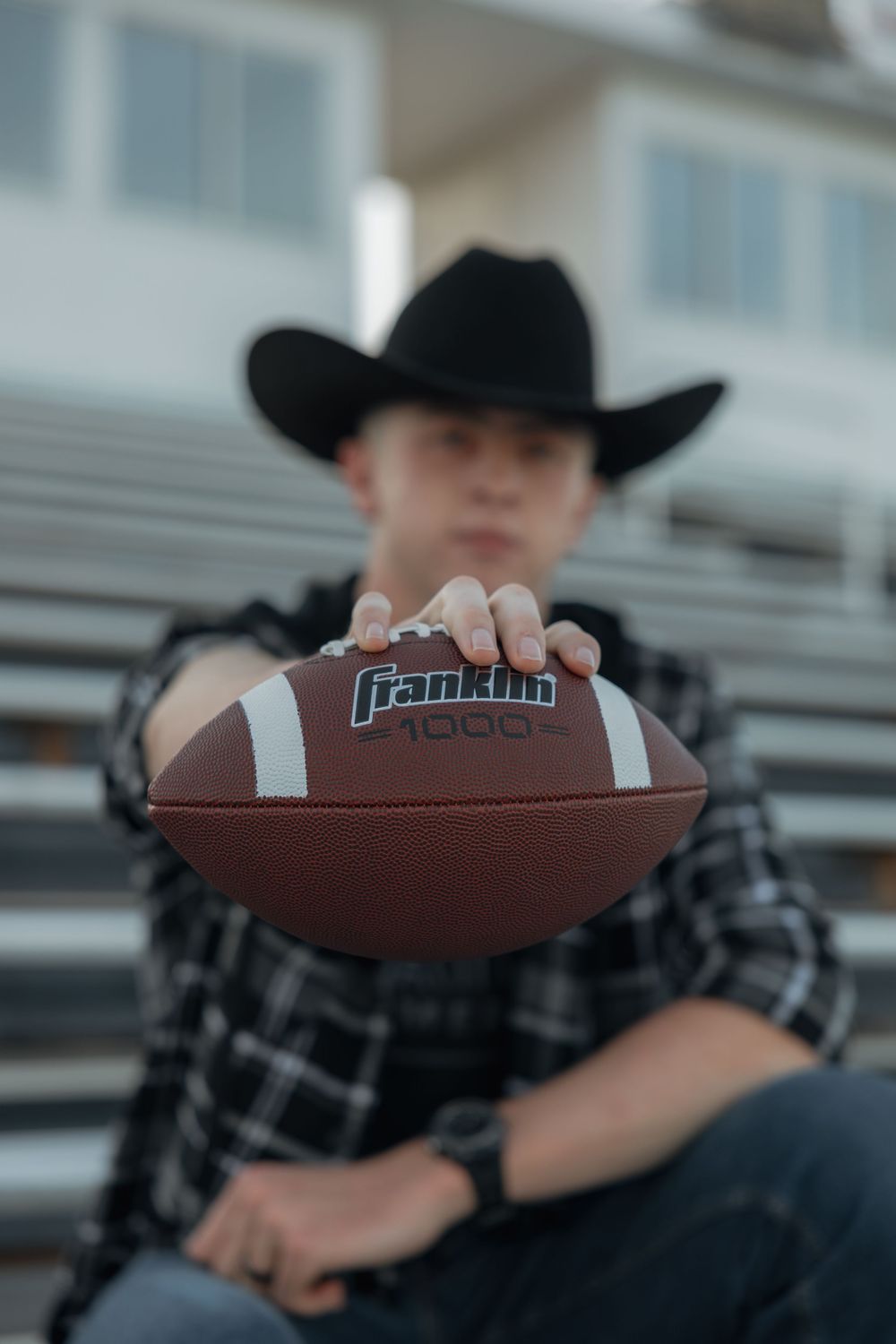 Young man in cowboy hat holding Franklin football toward camera in stadium bleachers.