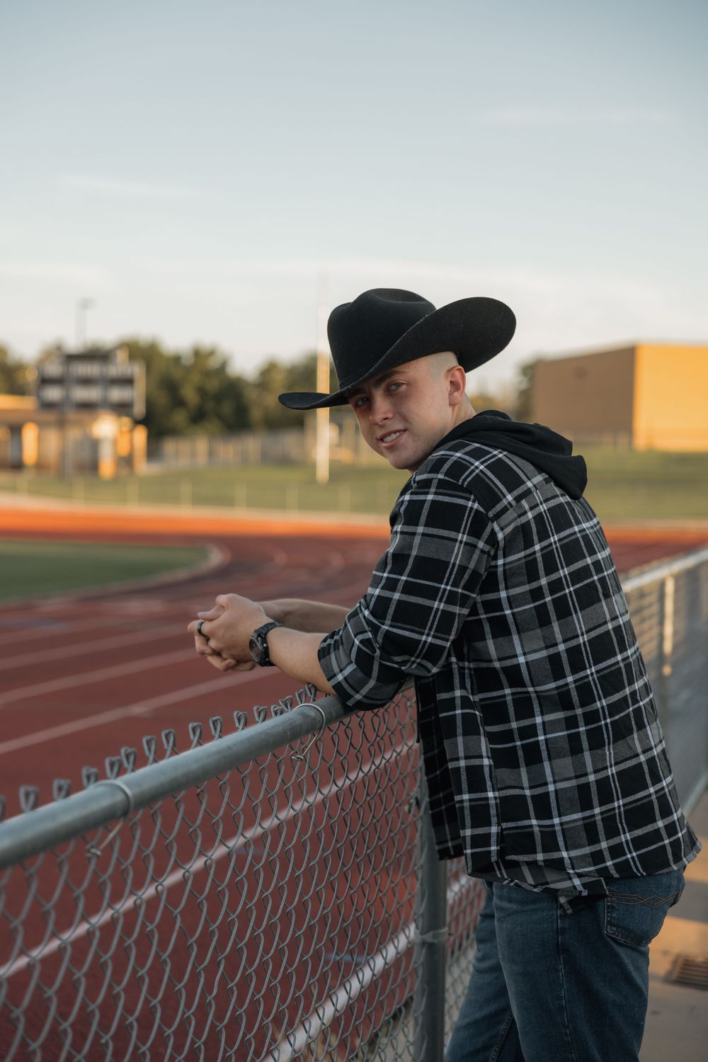 Young man in black cowboy hat leaning on chain-link fence at athletic track stadium.