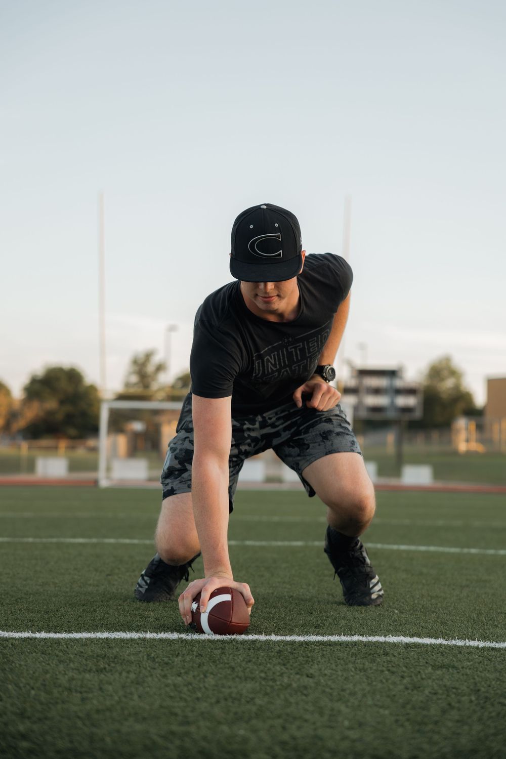 Young man in black cap and camo shorts placing football on field at sunset.