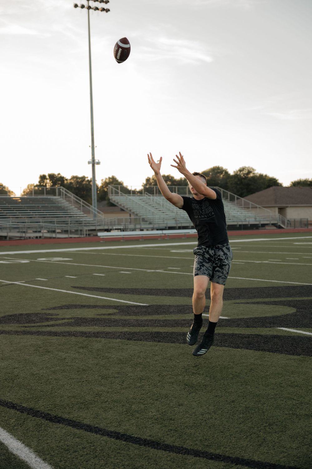 Young man reaching up to catch football on green turf field at dusk.