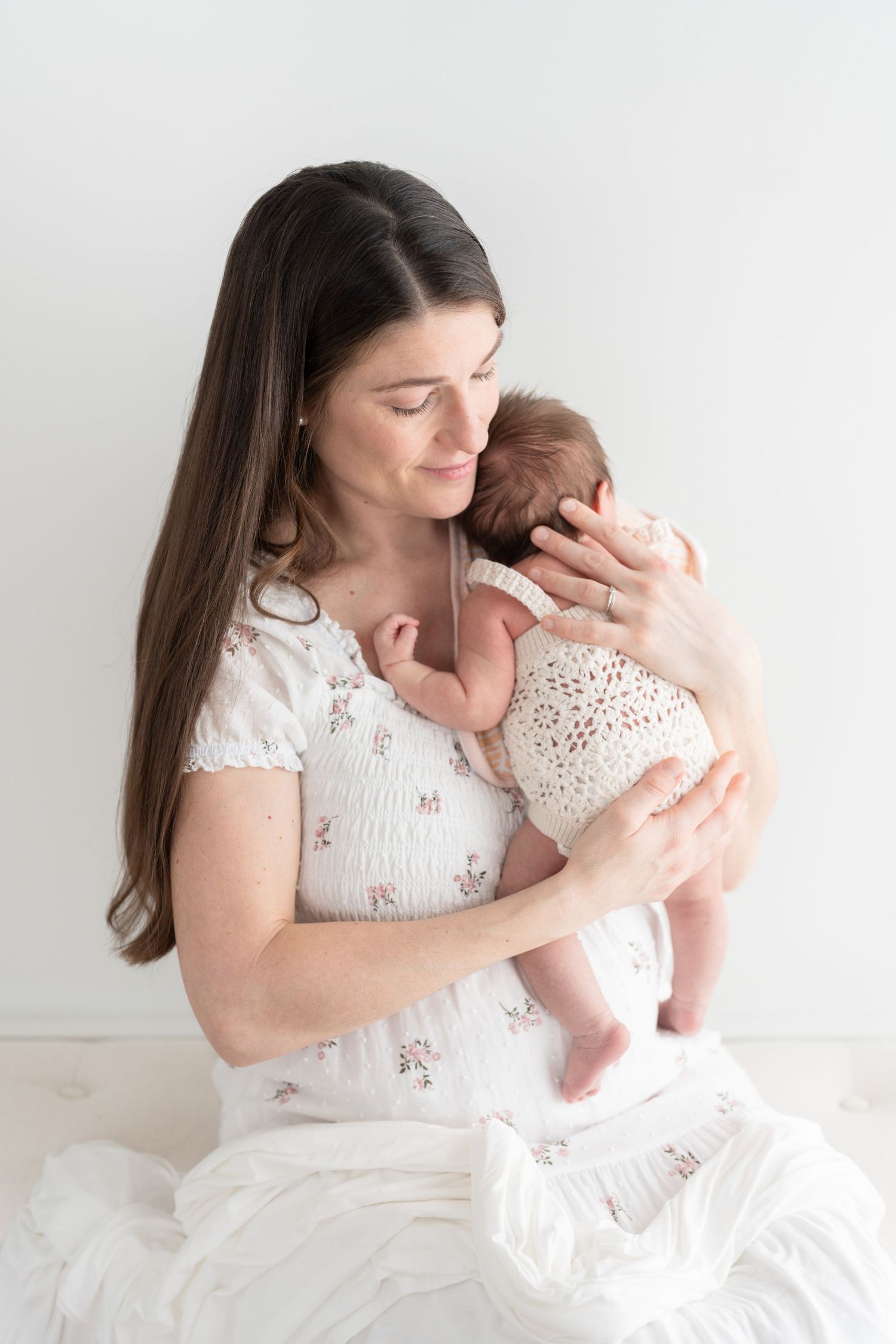Nouvelle maman vêtue d’une robe blanche a fleurs délicates tenant son bébé contre son épaule avec amour lors d’une séance nouveau-né studio