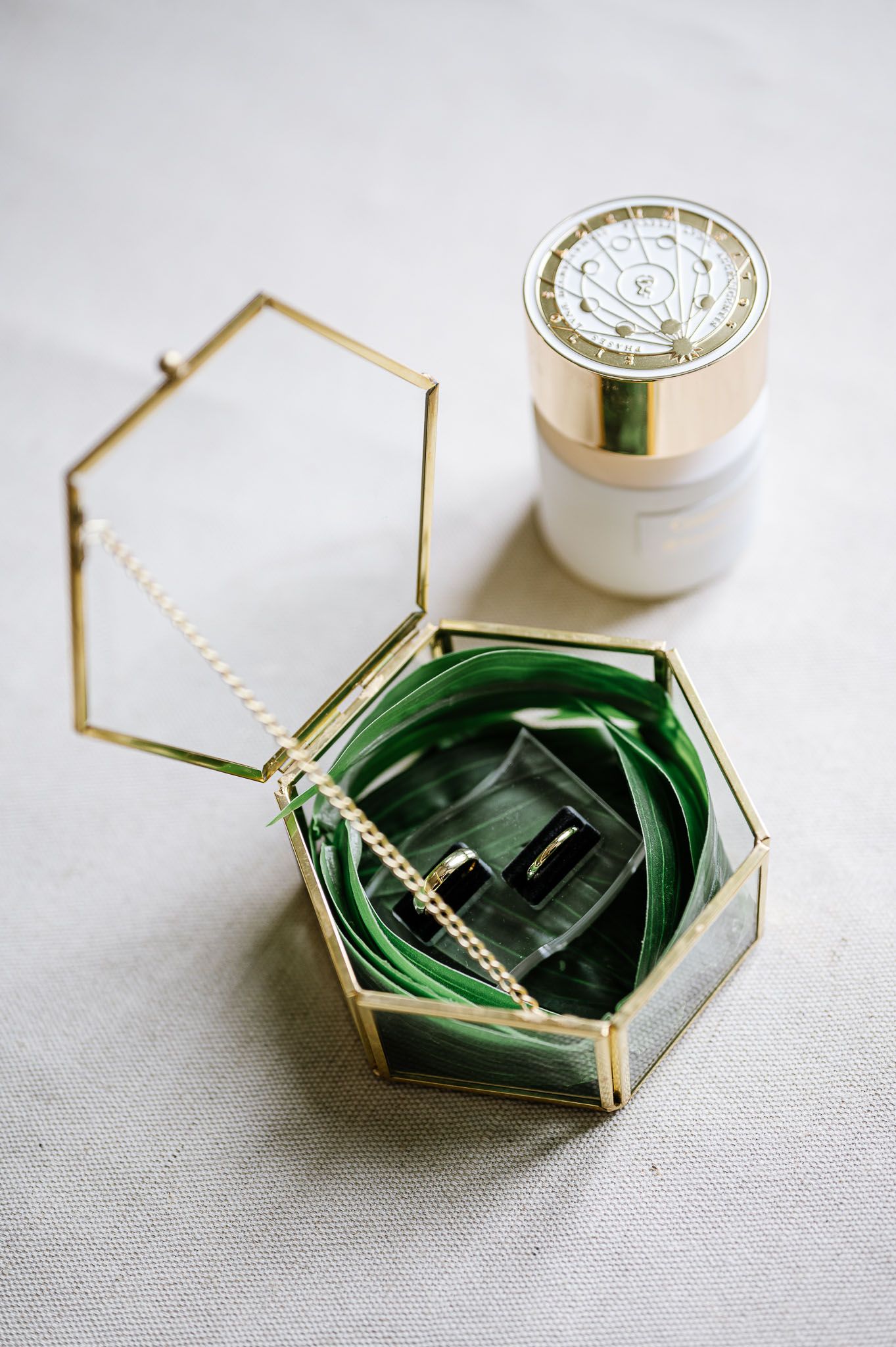 Gold geometric glass ring box with green velvet interior holding wedding bands, beside a white floral ring holder.