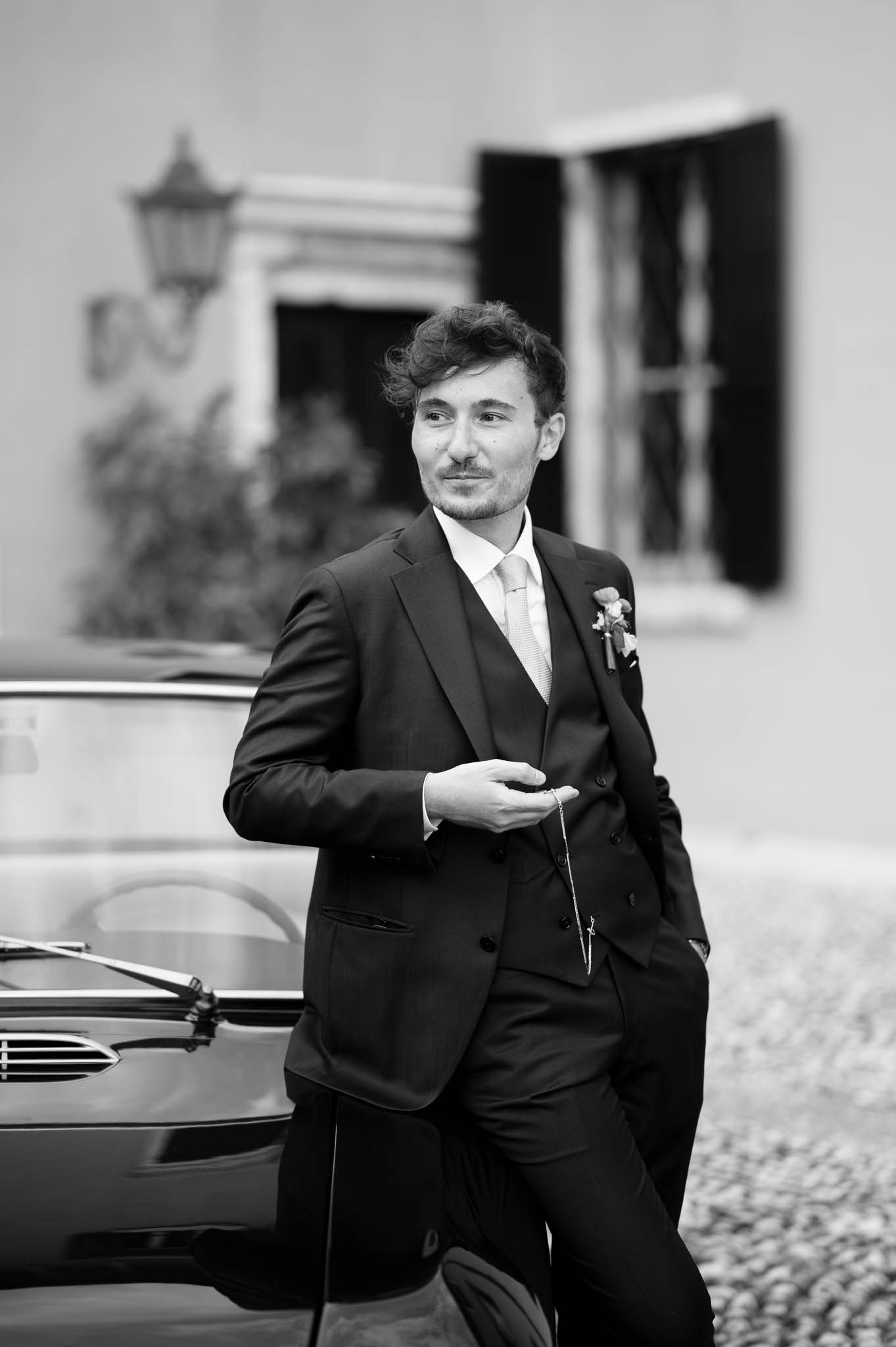 Elegant groom in black suit leaning against vintage car outside Villa della Torre in black and white wedding photo.