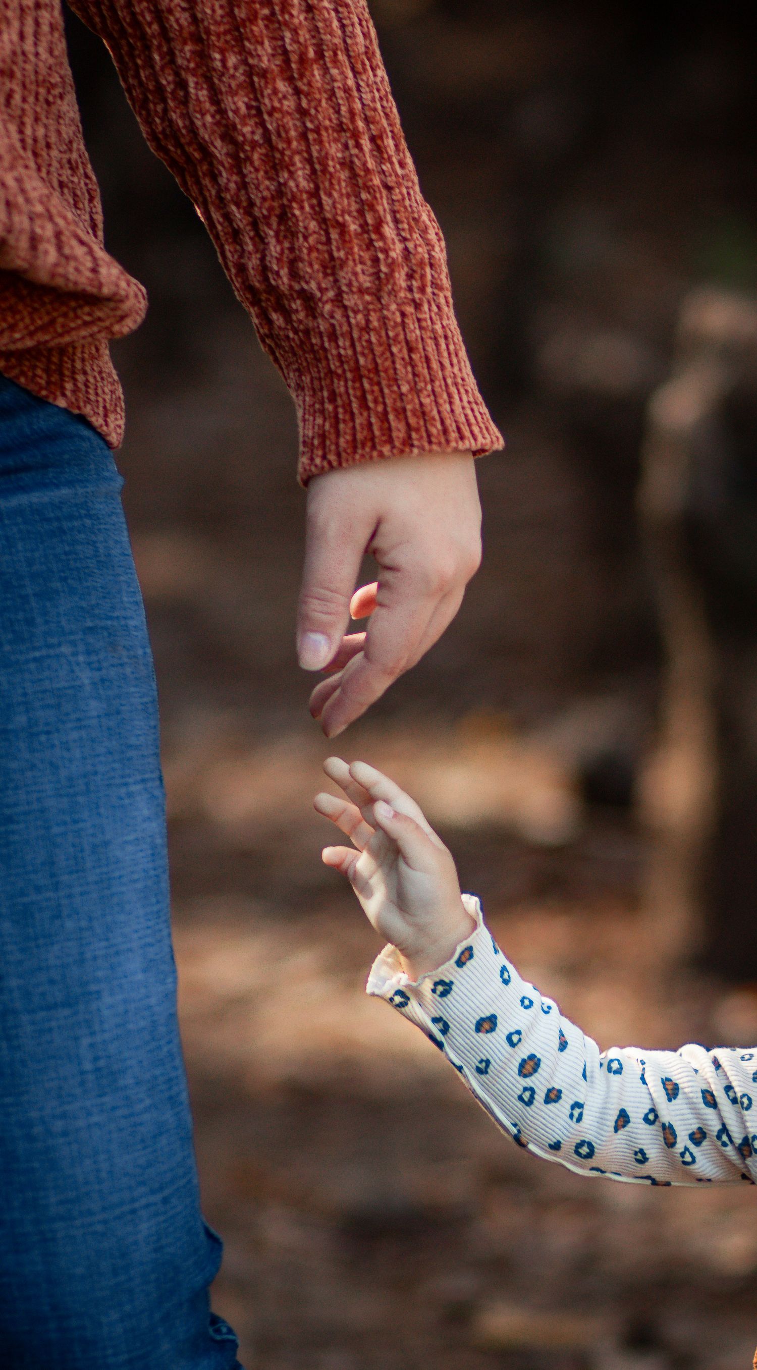 Adult in red sweater reaching down to hold a small child's hand outdoors in a blurred natural setting.