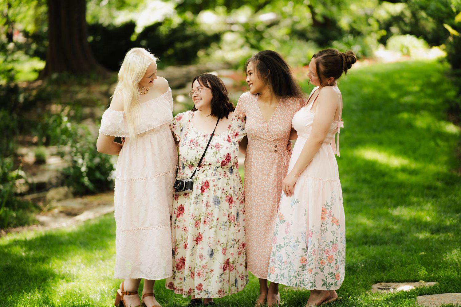 Four women in pastel floral dresses laughing together on lush green lawn in sunlight.