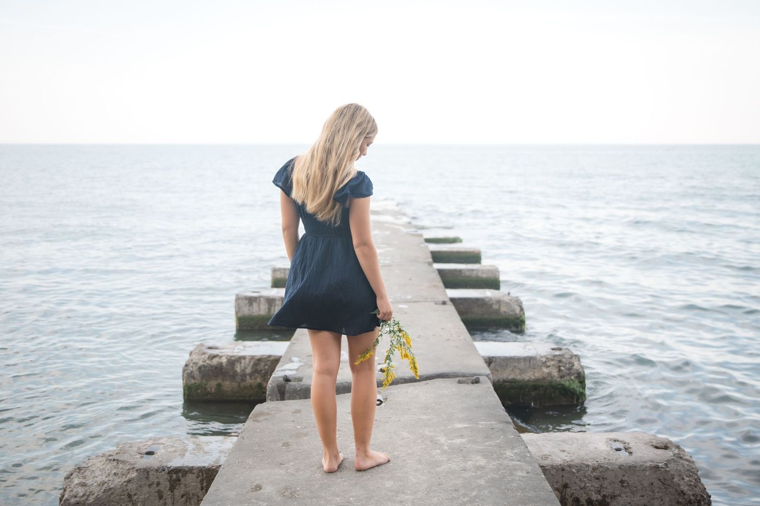 Blonde woman in navy dress standing barefoot on a stone pier, gazing at the calm ocean horizon.
