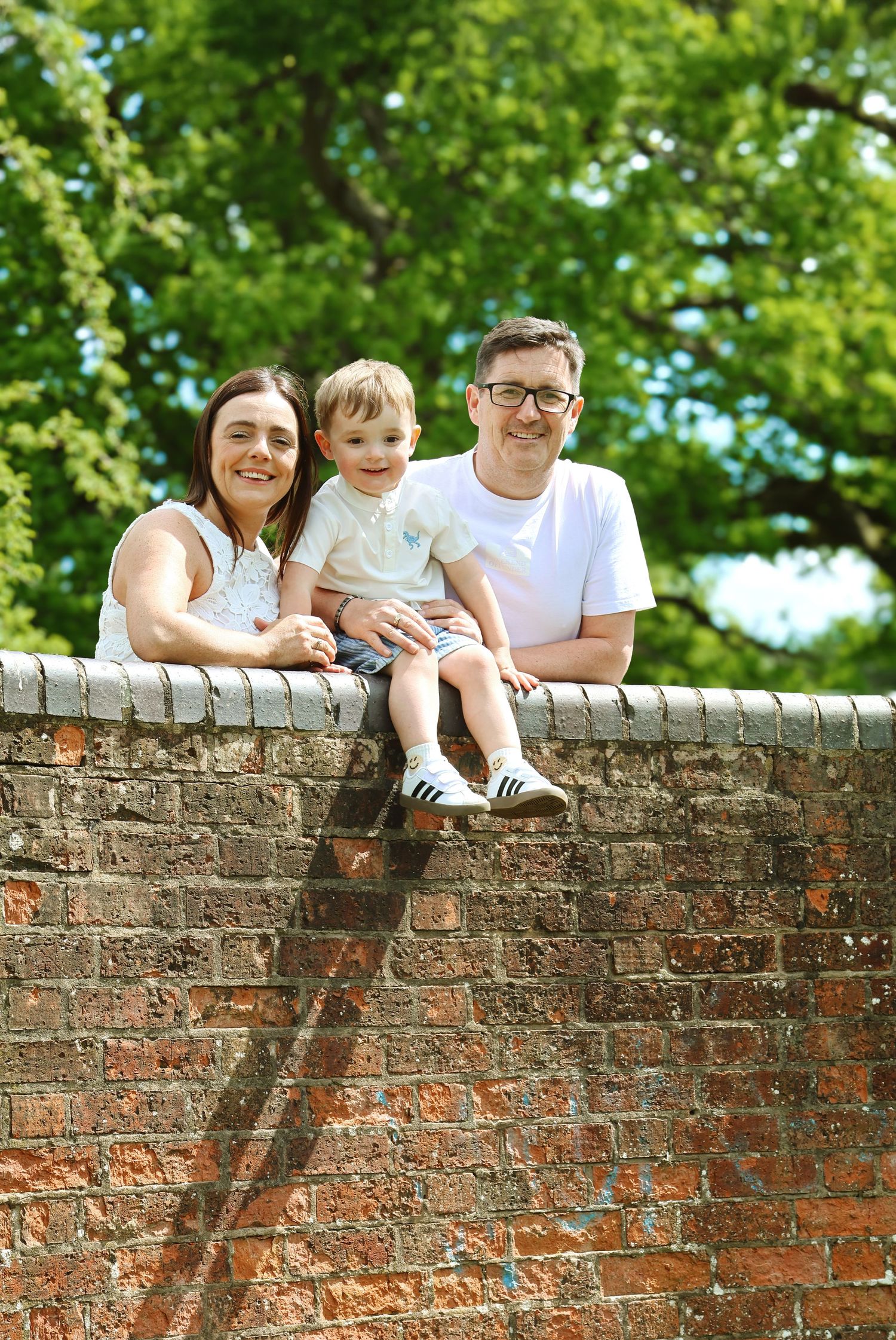 A happy family of three poses on a brick wall outdoors, with lush green trees in the background on a sunny day.