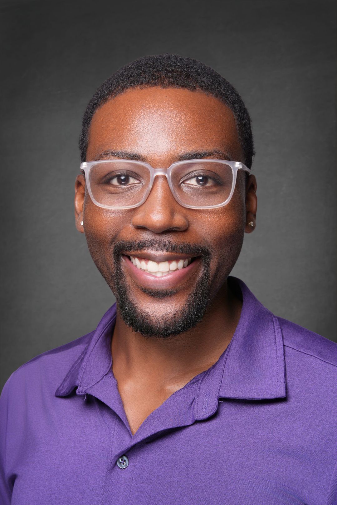 Smiling man with a trimmed beard and clear-framed glasses wearing a purple polo shirt in a professional headshot against a gray background.