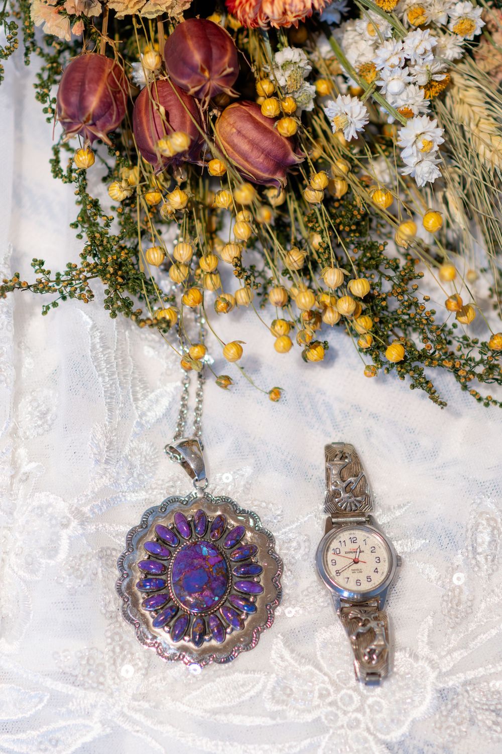 Silver Native American jewelry with purple turquoise pendant and watch on white lace beside dried floral bouquet.