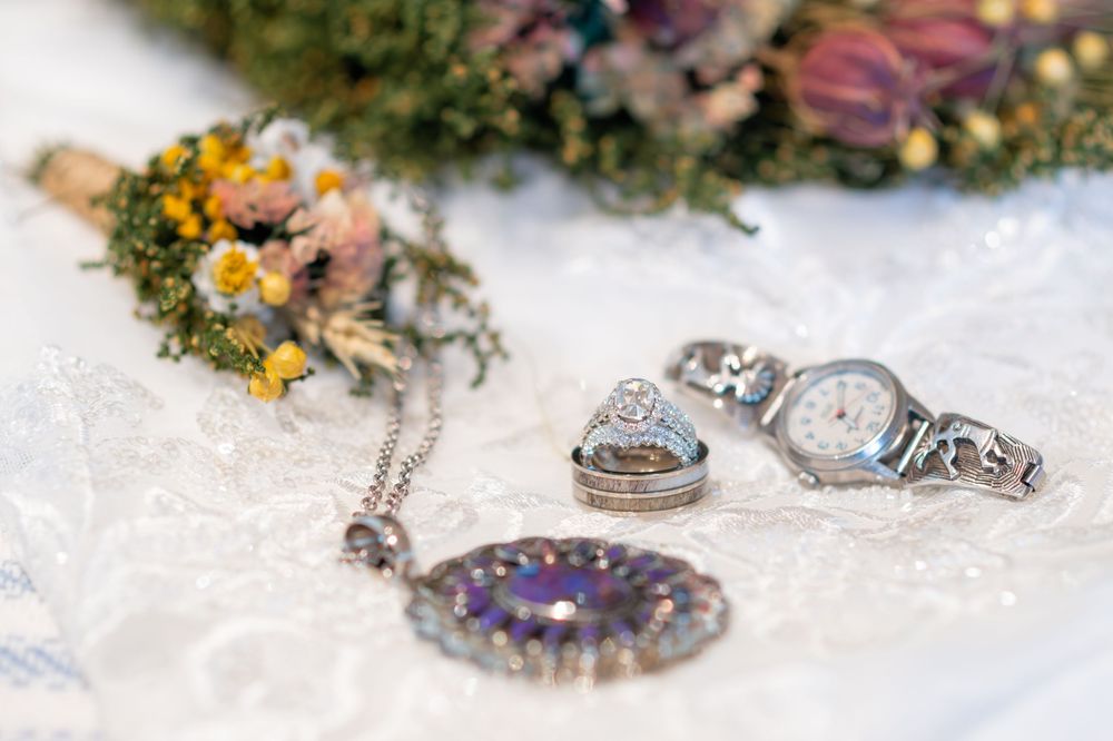 Wedding rings, silver watch, and purple gemstone pendant on white lace with dried flowers in background.