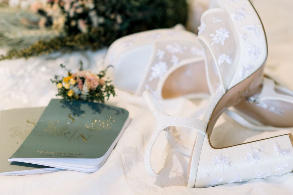 White lace bridal heels with floral embellishments beside wedding vow book and small bouquet.