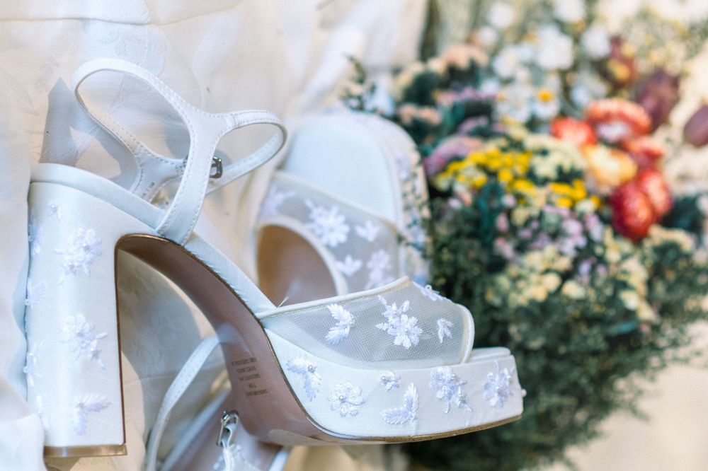 White platform bridal heels with floral appliques displayed in front of colorful wedding bouquet.