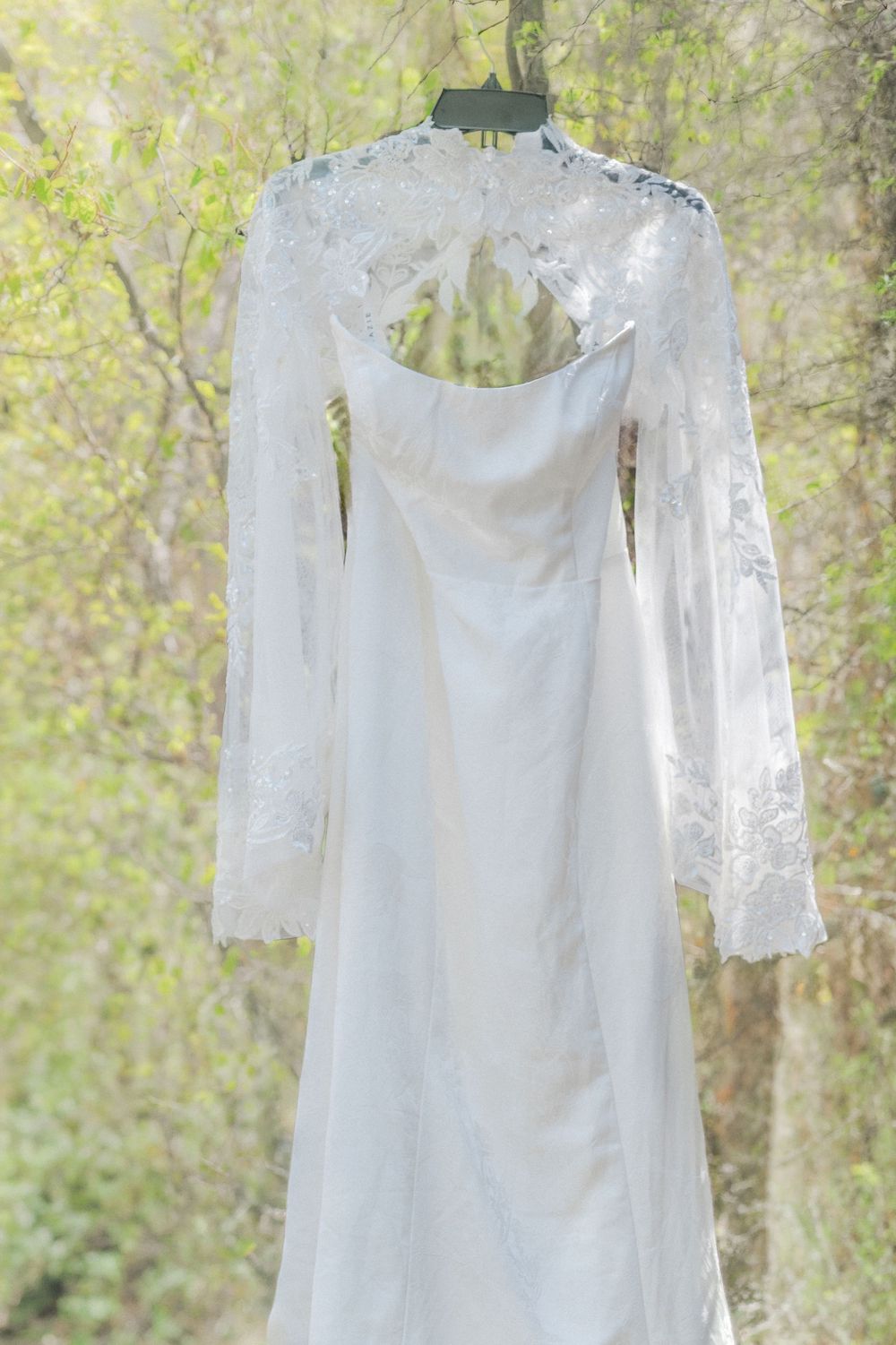 White lace long-sleeve wedding dress with open back hanging outdoors among green trees.