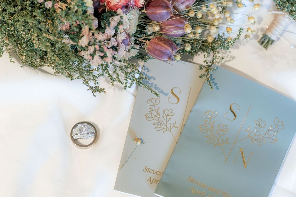 Wedding flat lay with sage green vow books, ring box, and colorful wildflower bouquet.