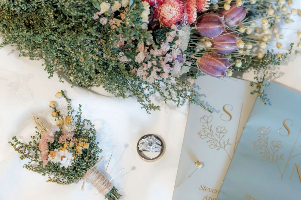 Wedding flat lay featuring wildflower bouquets, a ring box, and sage green monogram vow books.
