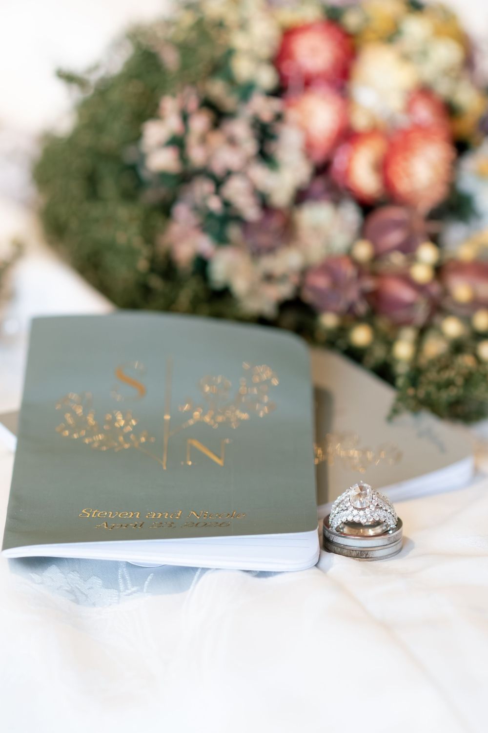 Wedding rings resting on sage green monogrammed vow books beside a dried floral bouquet.