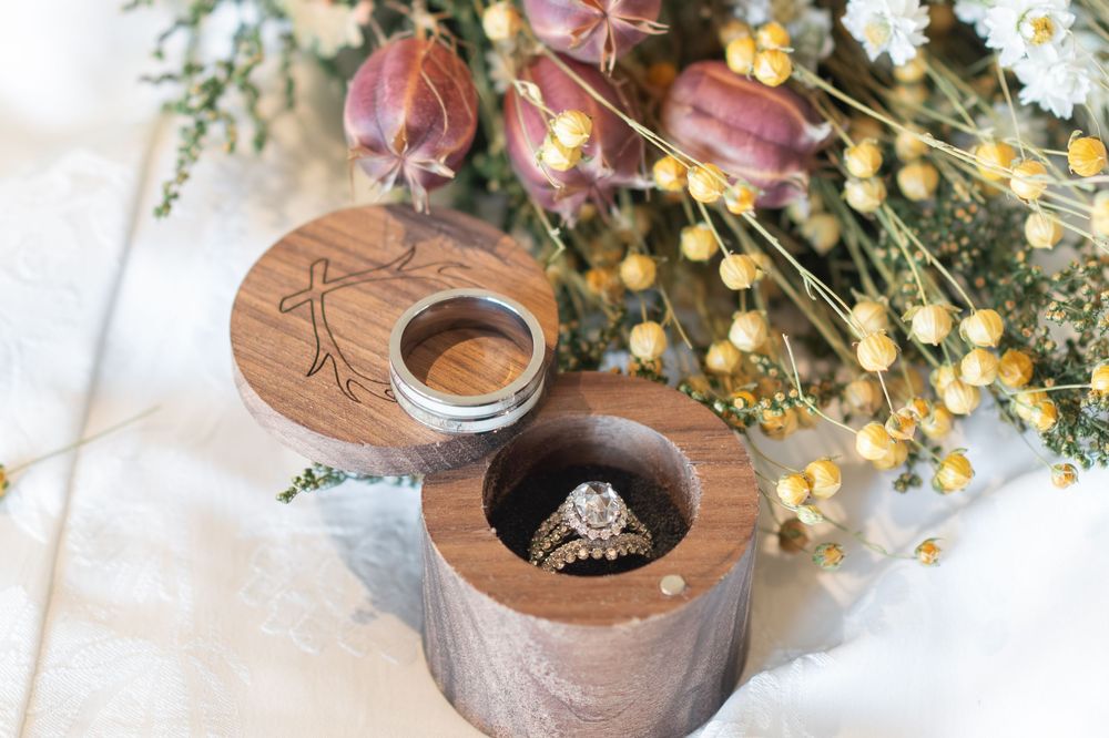 Diamond engagement ring and gold band in an engraved wooden ring box with dried wildflowers.