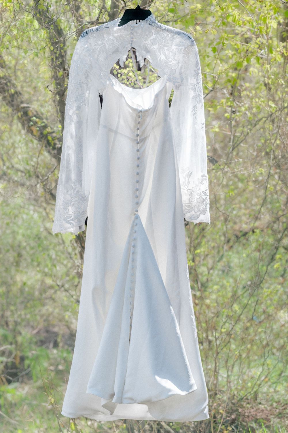 White lace wedding dress with long sleeves hanging on a hanger outdoors with green trees in background.