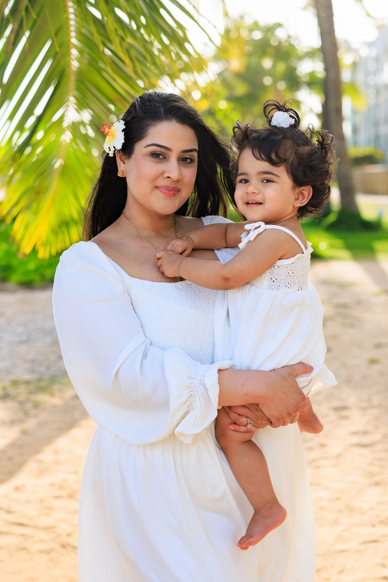 A woman in a white dress holds a toddler girl in white on a sunny beach with palm trees.
